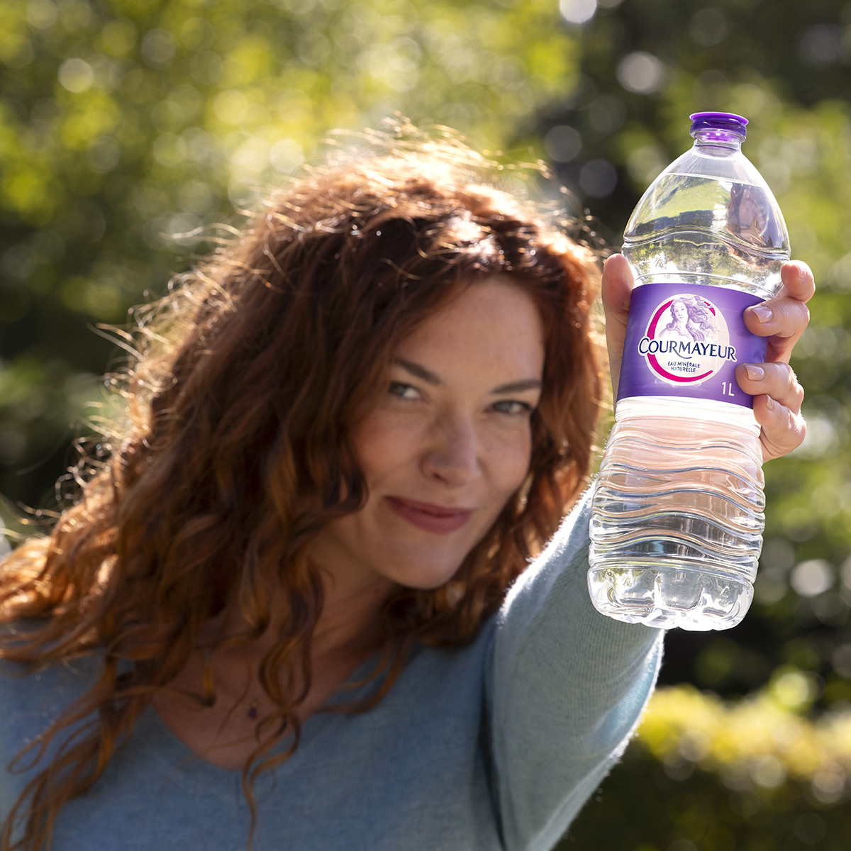 Une jeune femme rousse aux cheveux longs ressemblant à la Venus du logo tend devant elle une bouteille de Courmayeur.